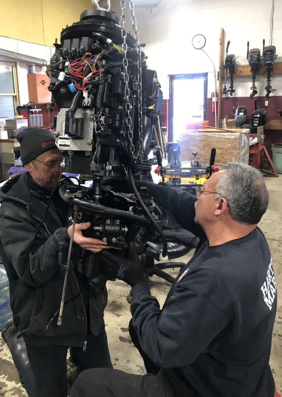Two technicians rebuilding an outboard powerhead lifted on a chain hoist, wiring exposed